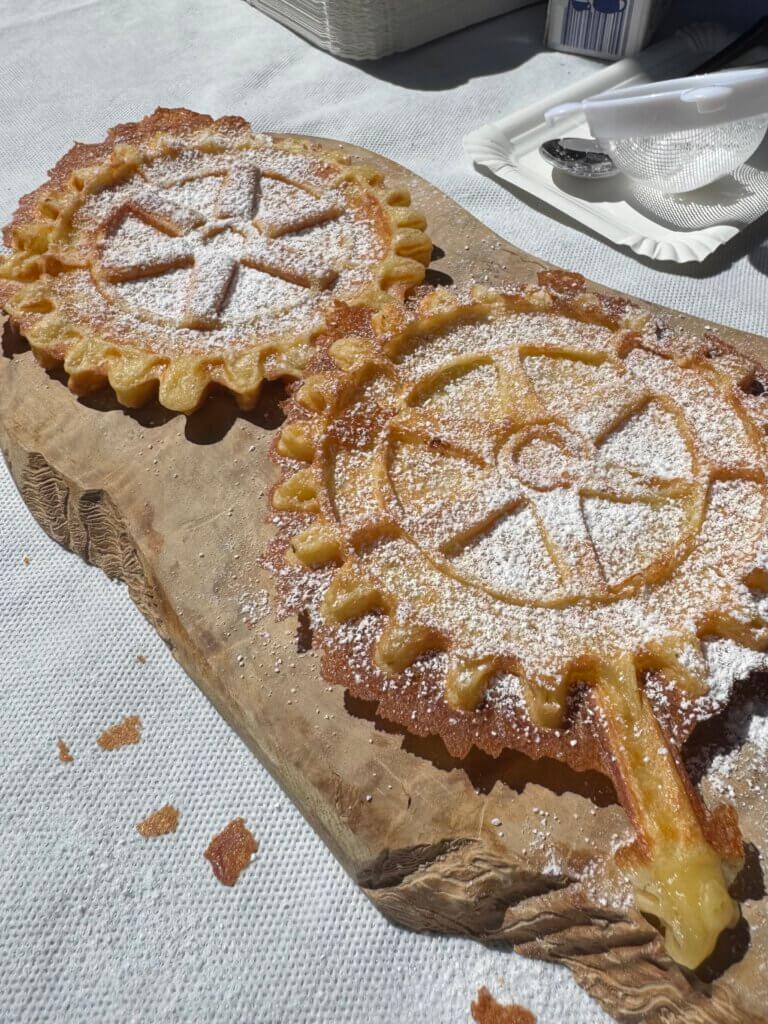 Zwei frisch gebackene Waffeln auf einem Holzbrett liegend und mit Puderzucker bestreut. Die Waffelform zeigt das Logo des Rotarier Clubs.