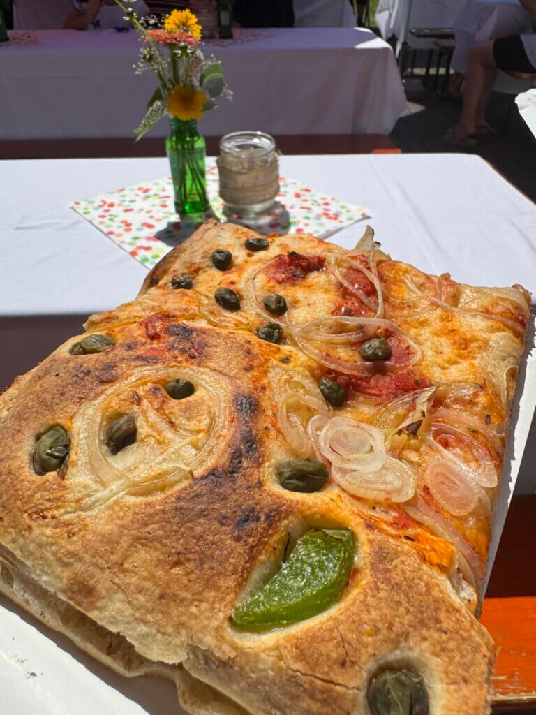 Ein qudratisches Stück Pizza, belegt mit zwiebeln, grünen peperoni und Oliven.