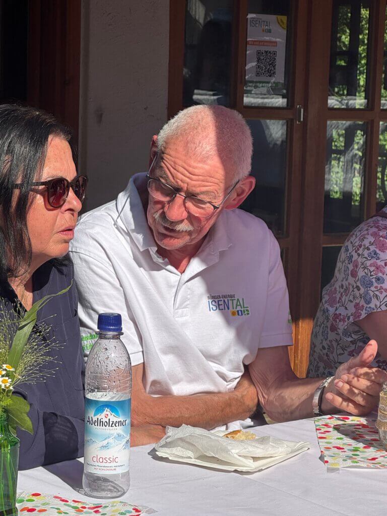Älterer Mann im Gespräch mit mit einer dunkelhaarigen Frau mit Sonnenbrille, am Tisch sitzend.