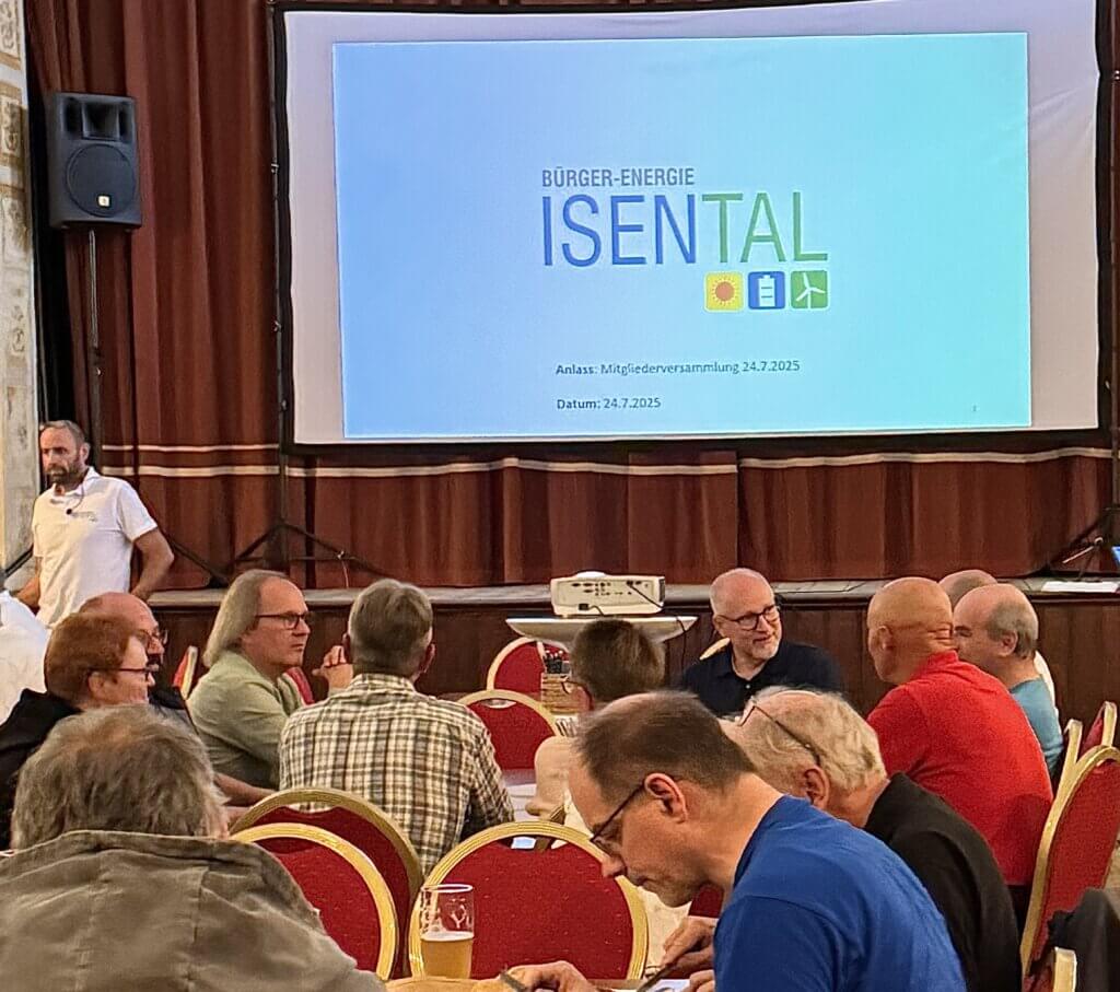 Mehrere Personen sitzen an Tischen mit roten Stühlen in einem Veranstaltungsraum und unterhalten sich. Im Hintergrund ist eine Leinwand mit einer Projektion zu sehen. Die Projektion zeigt den Schriftzug ‚BÜRGER-ENERGIE ISENTAL‘ sowie den Anlass ‚Mitgliederversammlung 24.7.2025‘.
