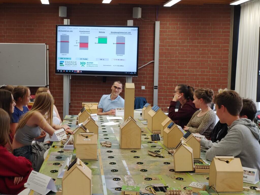 Ein Gruppenraum mit einem großen rechteckigen Tisch, auf dem mehrere kleine Holzmodelle von Häusern stehen. Jedes Modell ist mit kleinen Solarpanels ausgestattet. Um den Tisch sitzen mehrere Personen, die sich offenbar an einer interaktiven Übung beteiligen. Im Hintergrund hängt ein großer Bildschirm an einer roten Backsteinwand, der Diagramme und Balkengrafiken zeigt. Darunter sind Logos von Organisationen wie „Energieagentur“ und „Bayerische Landesanstalt für Wald und Forstwirtschaft“ sichtbar. Die Atmosphäre wirkt wie ein Workshop oder eine Schulung zum Thema erneuerbare Energien.