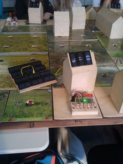 Nahaufnahme eines Holzmodells in Form eines Hauses, das auf einer Tischfläche mit Landschaftsmotiven steht. Auf dem Dach des Modells sind mehrere kleine Solarpanels angebracht. Daneben sind Kabel und eine kleine Einheit mit Schaltern und LEDs zu sehen, die offenbar den Energiefluss simulieren. Weitere ähnliche Modelle sind im Hintergrund erkennbar. Die Tischfläche zeigt grüne Wiesen und Felder, was den Bezug zu nachhaltiger Energie und Natur verdeutlicht.