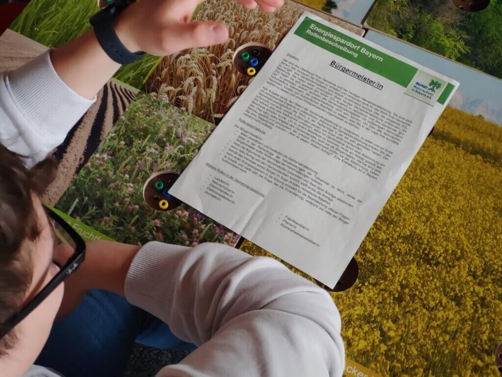 Ein Tisch mit einer Spielfläche, die landwirtschaftliche Felder und Wiesen darstellt. Darauf liegt ein Informationsblatt mit dem Titel „Energiespardorf Bayern – Rollenbeschreibung Bürgermeister/in“. Das Blatt enthält Textabschnitte zu Aufgaben und Zielen dieser Rolle. Eine Person sitzt davor und hält die Hände leicht über das Papier, als würde sie sich auf die Inhalte konzentrieren. Die Umgebung deutet auf ein Planspiel oder eine Simulation zum Thema Energie und Kommunalpolitik hin.