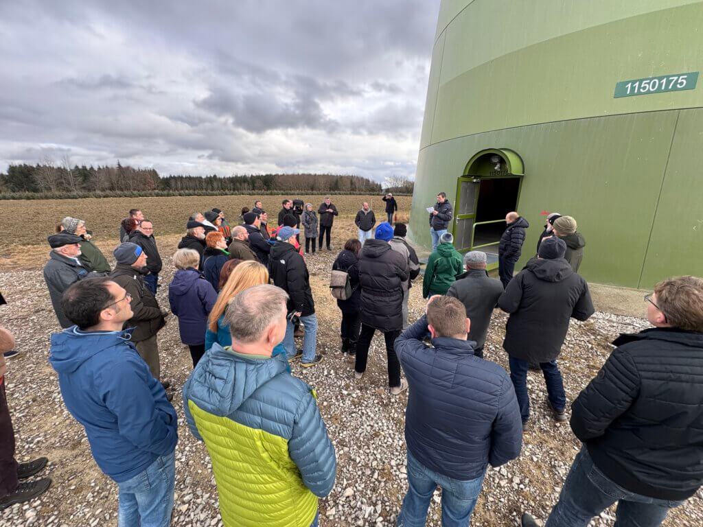 Gruppe von Menschen steht vor dem Eingang eines großen grünen Windradturms auf einem Feld unter bewölktem Himmel.