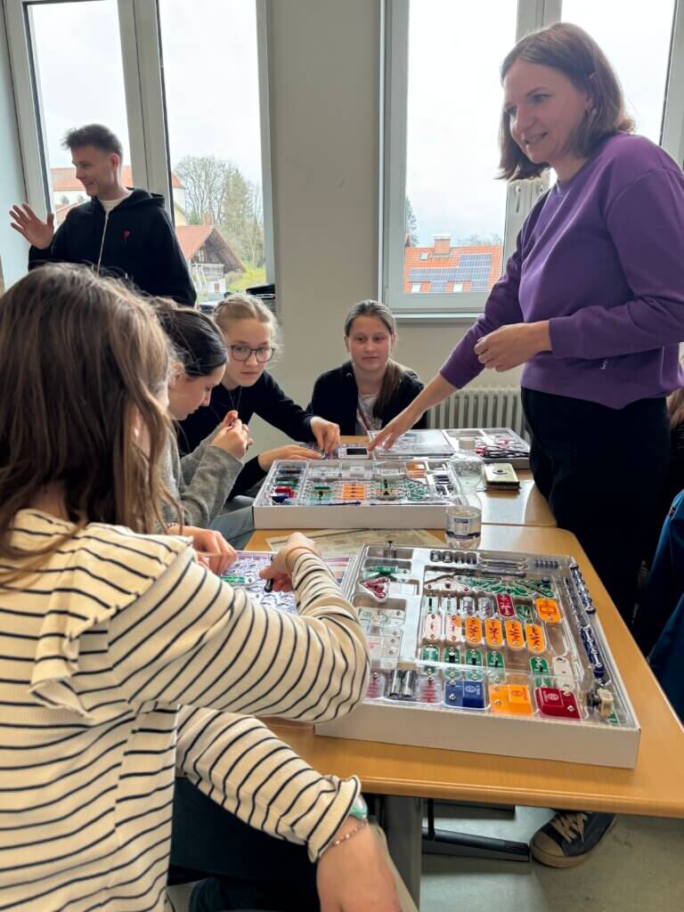 Mehrere Mädchen sitzen an einem Tisch und arbeiten mit elektronischen Bauteilen, eine Frau steht daneben und erklärt etwas, im Hintergrund Fenster mit Blick auf Häuser.