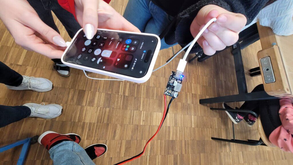 Hand hält Smartphone mit angeschlossenem Kabel zu einer kleinen Platine auf Holzboden mit mehreren Personen und Stuhlbeinen im Hintergrund