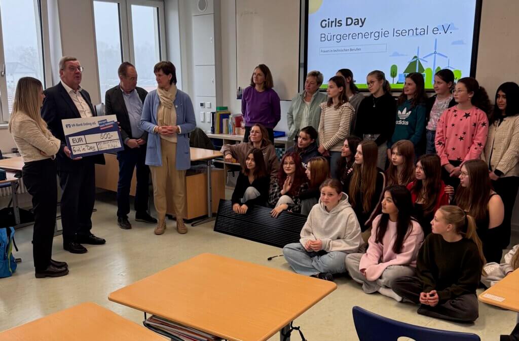 Gruppe von Mädchen und Frauen in einem Klassenraum, einige sitzen auf dem Boden, andere stehen, links übergibt ein Mann mit Anzug einen großen symbolischen Scheck an eine Frau, im Hintergrund Bildschirm mit Text 'Girls Day Bürgerenergie Isental e.V.'