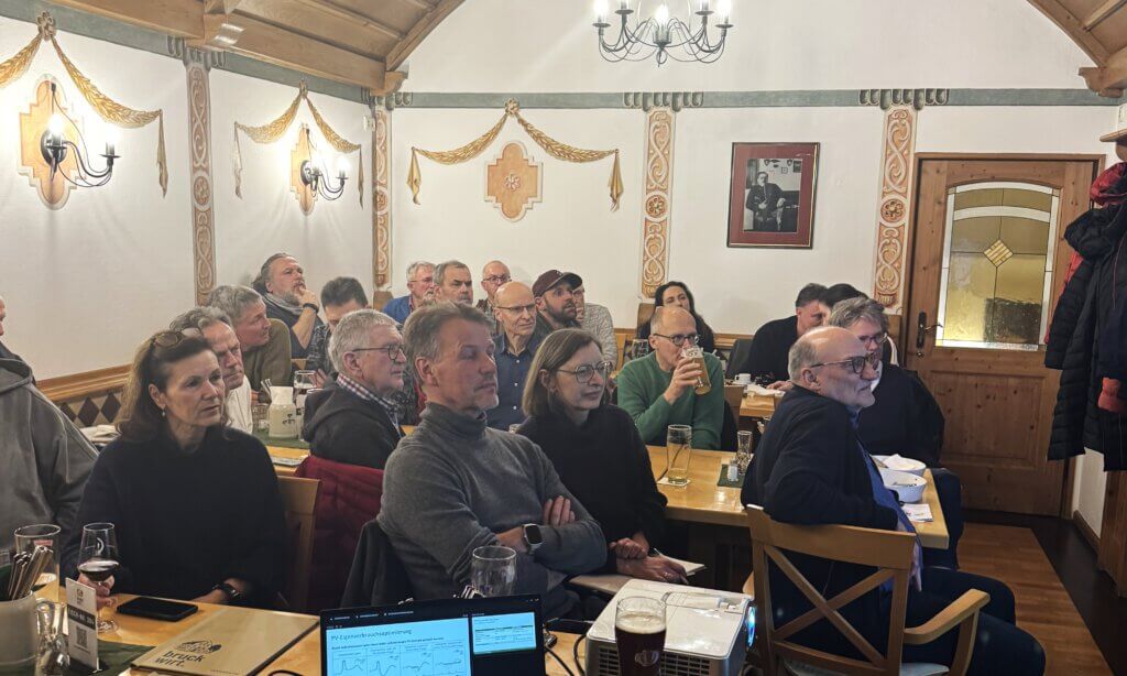 Mehrere Personen sitzen in einem rustikal eingerichteten Raum mit Holzbalkendecke und Wandlampen und hören einer Informationsveranstaltung zum BiDirektionalem Laden zu.